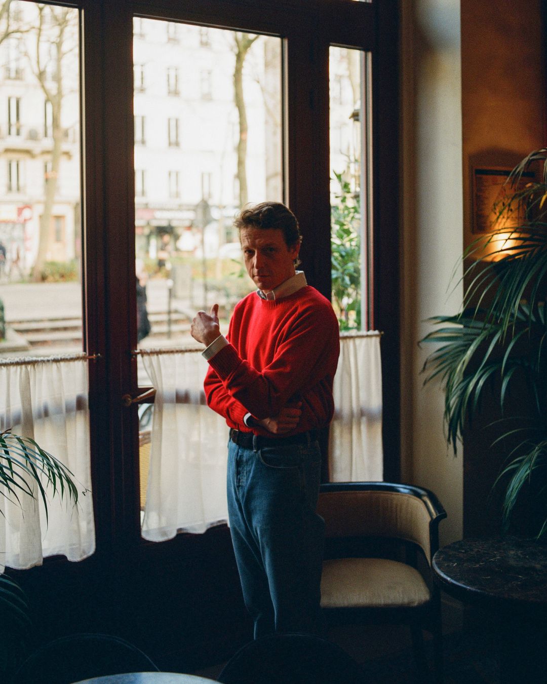 Man in a red sweater standing by a large window with a cityscape view.