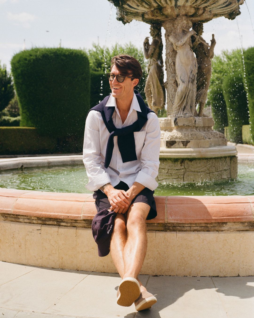 SS25: The Florence Edition: Person sitting on a fountain ledge in an outdoor setting with statues and greenery.