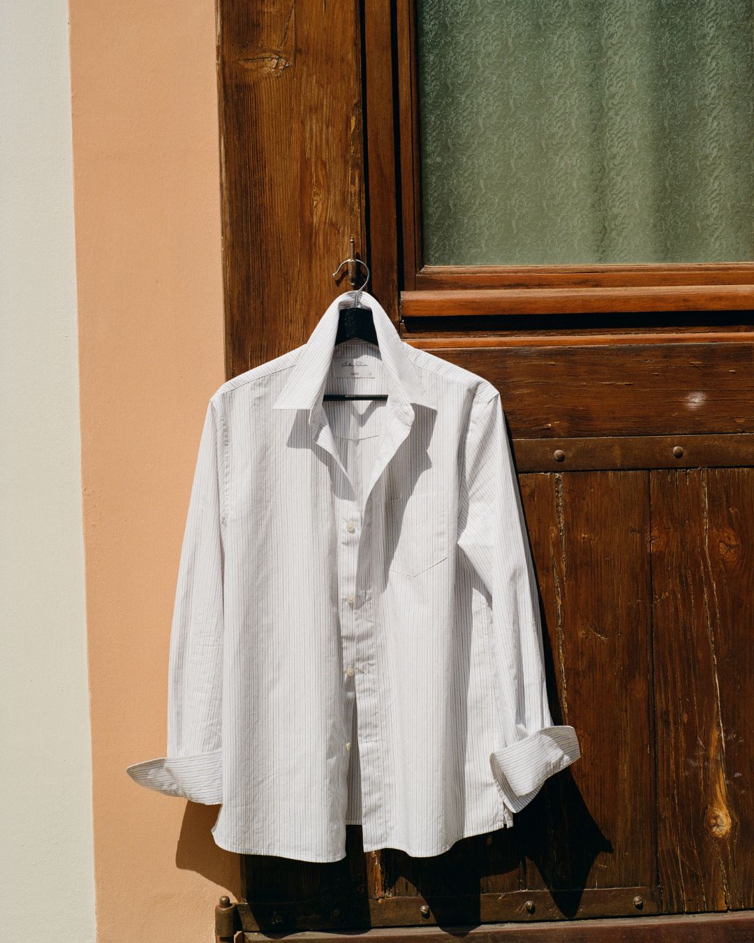 SS25_The_Florence_Edition_7: White shirt with a logo on a hanger against a wooden door.